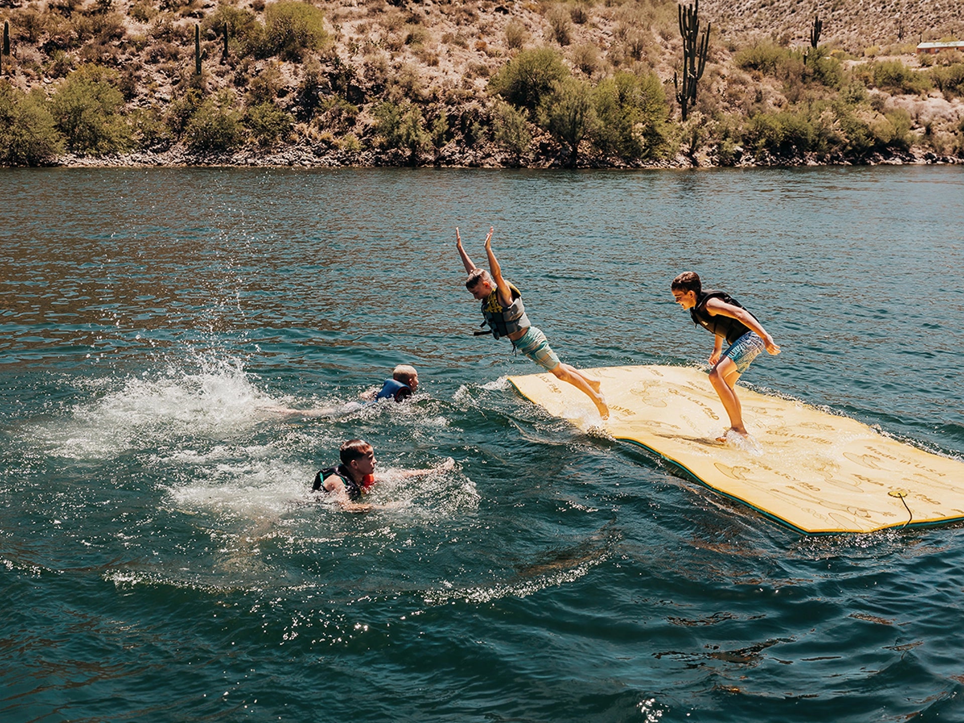 Four people enjoying a day at the lake, with one person on an Aqua Lily Pad and two others in the water.